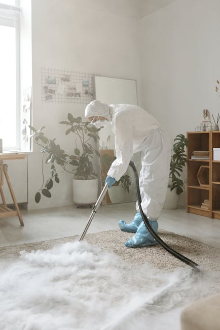 A person dressed in full protective clothing, including a white coverall, gloves, mask, and shoe covers, is performing deep cleaning on a carpet using a professional carpet cleaning machine with a hos