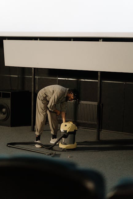 A person dressed in full protective clothing, including a white coverall, gloves, mask, and shoe covers, is performing deep cleaning on a carpet using a professional carpet cleaning machine with a hos